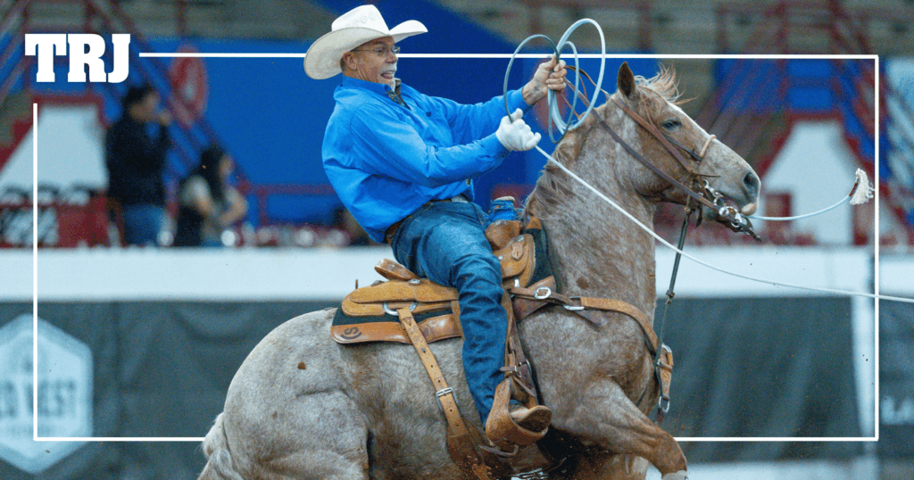 Header roper in a blue shirt and white cowboy hat swings his loop aboard a red roan horse in the arena