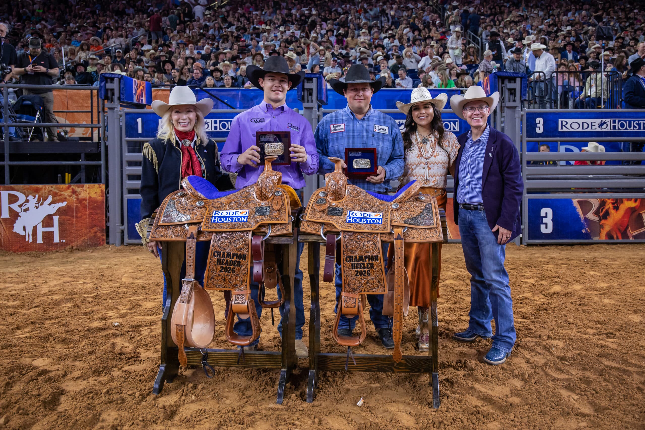 Rodeo winners posing with saddles in the arena surrounded by spectators and officials