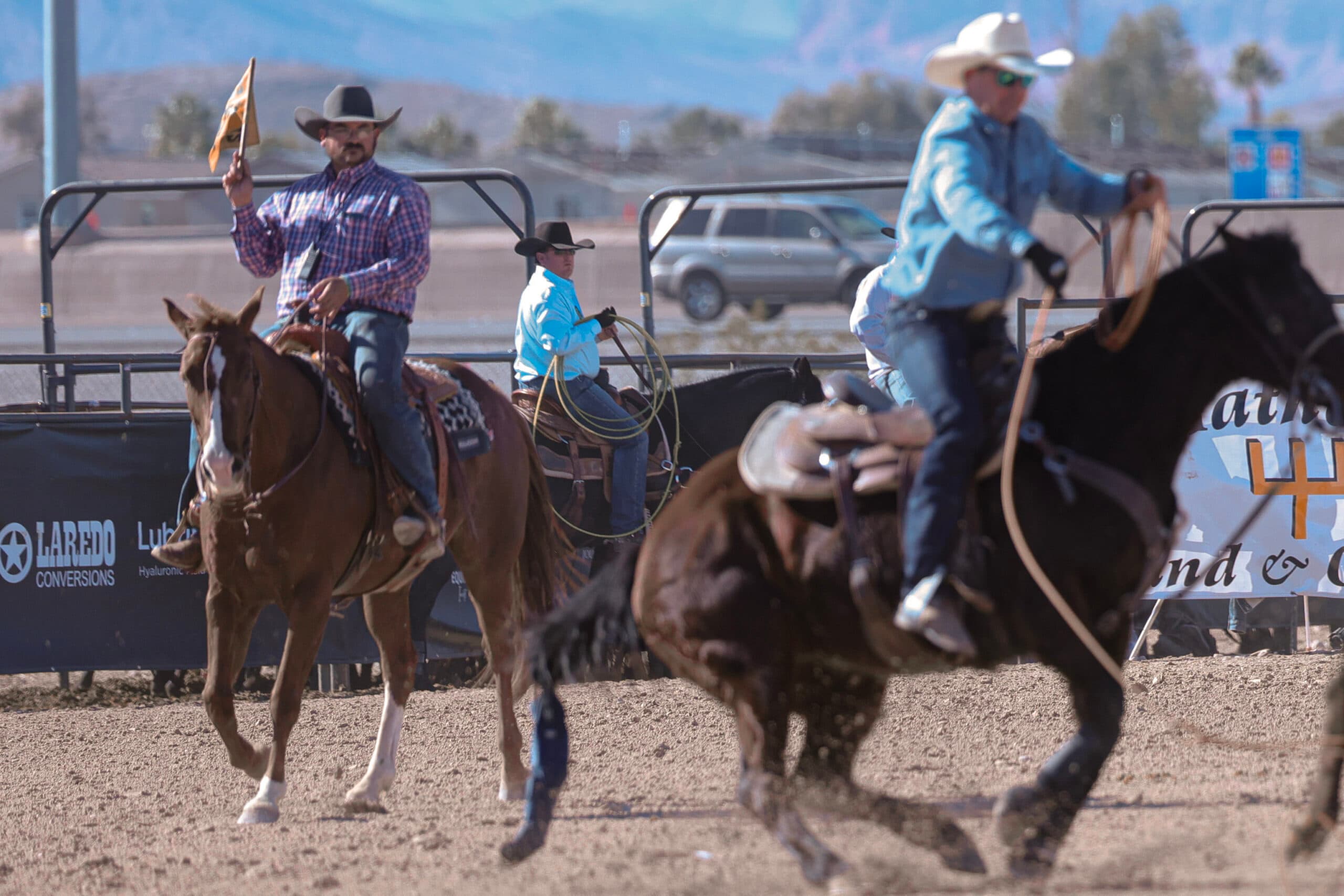 Flagman on a sorrel horse raises a yellow flag as a header on a dark horse drags a steer across the arena dirt