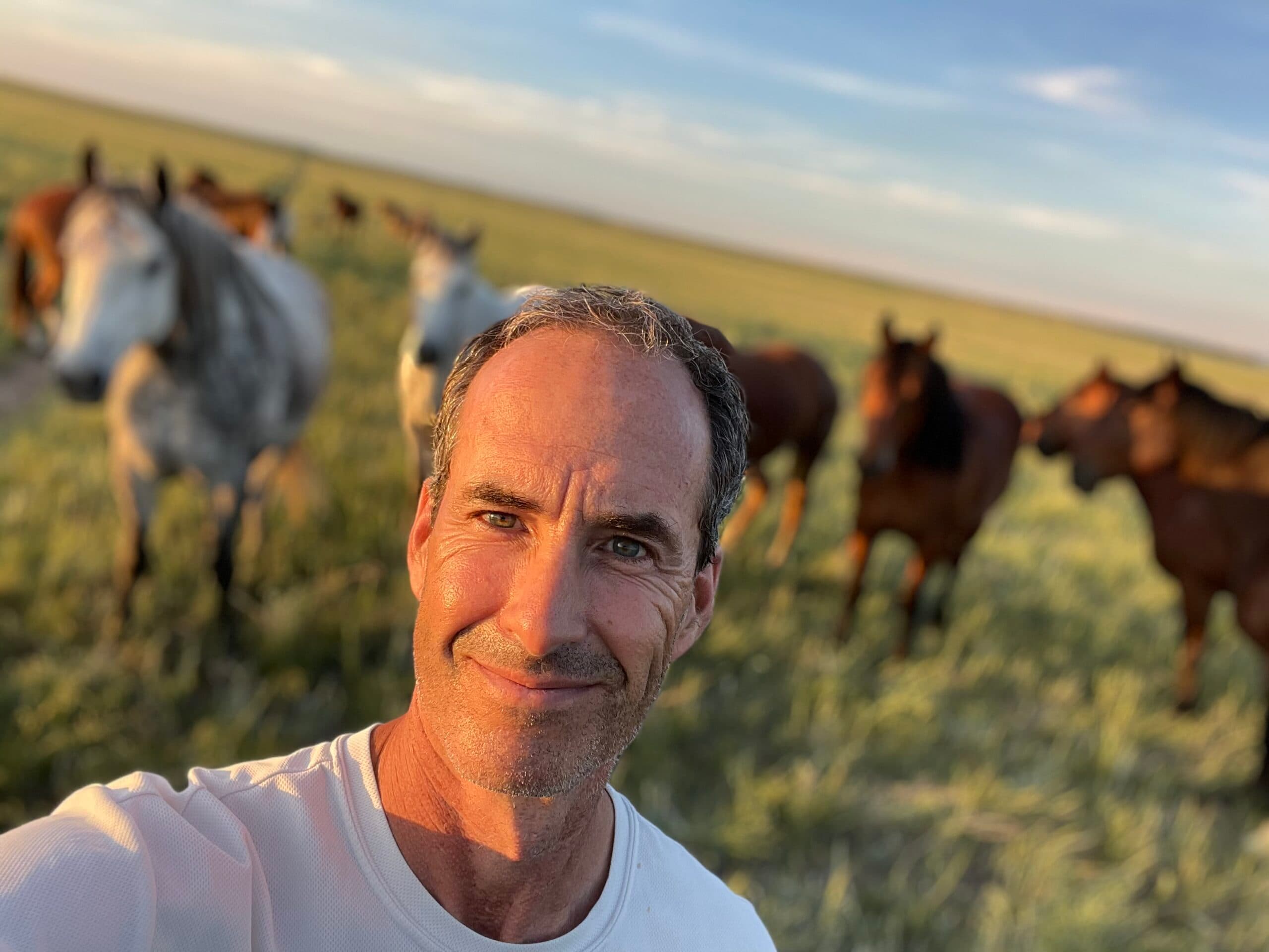 Man in white shirt smiles at camera with herd of horses grazing in pasture behind him