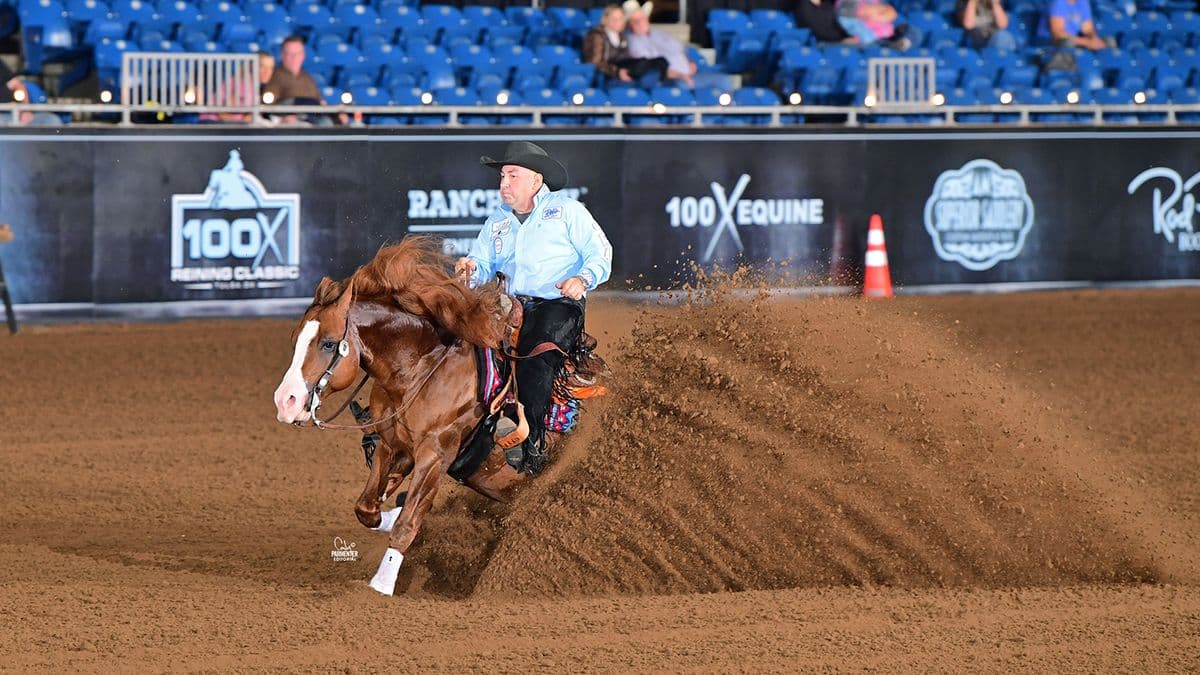 A reining horse and rider perform a sliding stop in an indoor arena, dirt flying as the horse digs into the ground with the crowd watching from the stands
