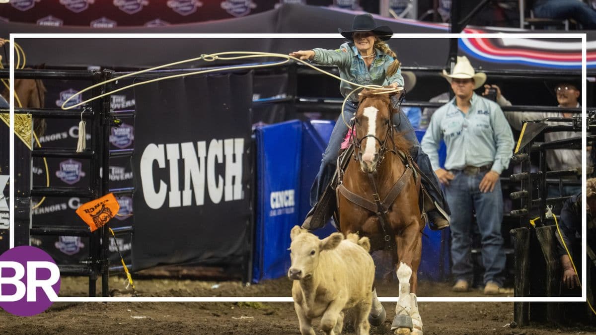 Cowgirl on horseback swings a lasso while herding a white calf during a ranch sorting competition at an indoor rodeo arena