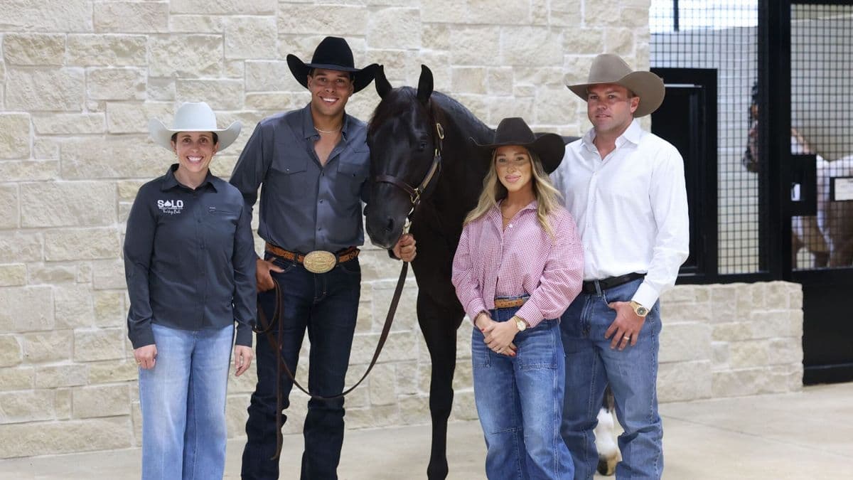 Five people in Western attire pose with a black horse in front of a stone barn