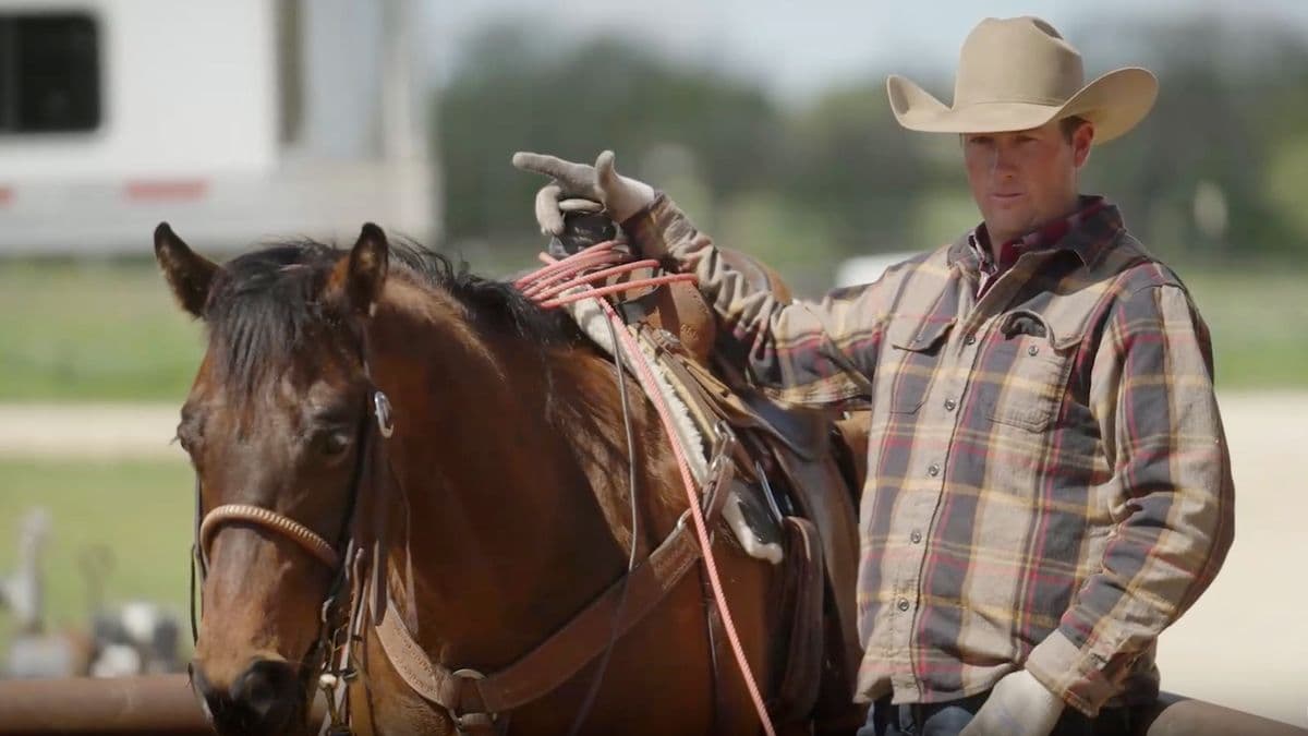Cowboy in plaid shirt and tan Stetson holds saddled brown horse's reins while standing at a rodeo grounds with horse trailer visible behind him