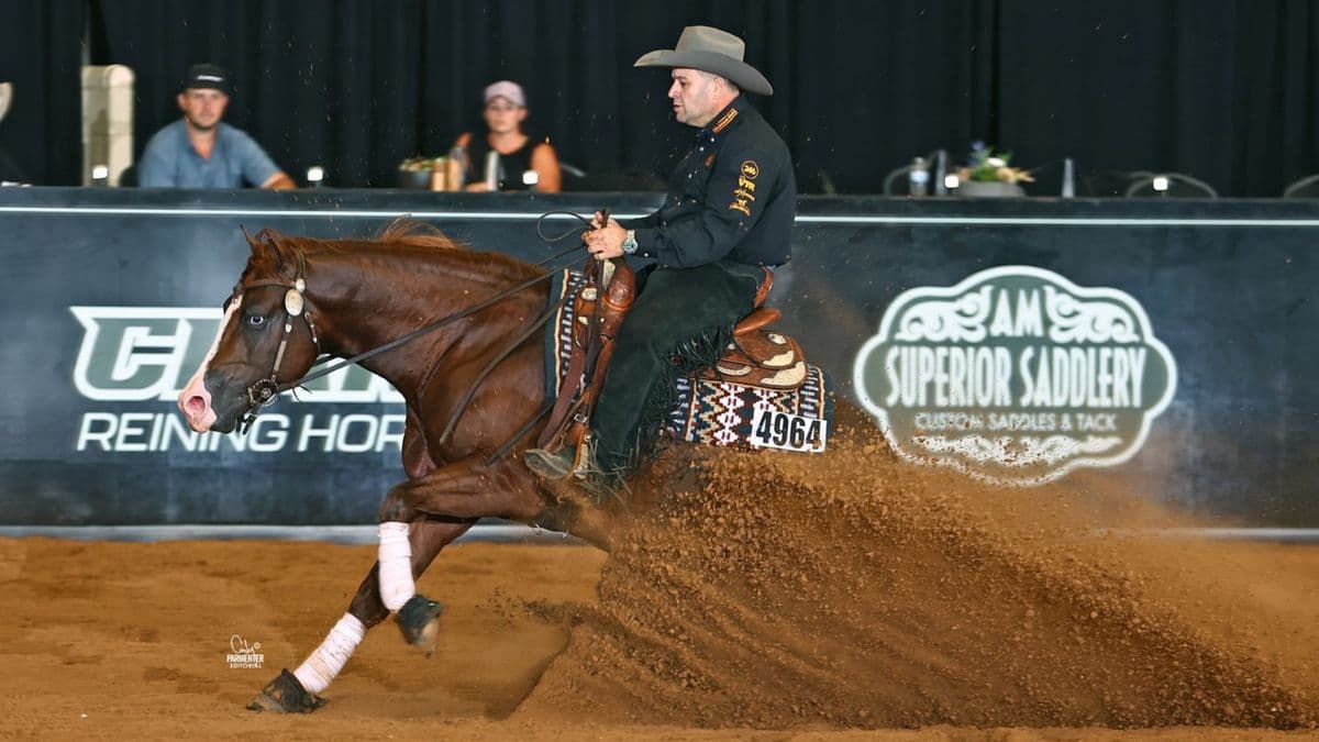 Reiner in black shirt and chaps slides his chestnut horse to a stop, spraying dirt in front of arena banners