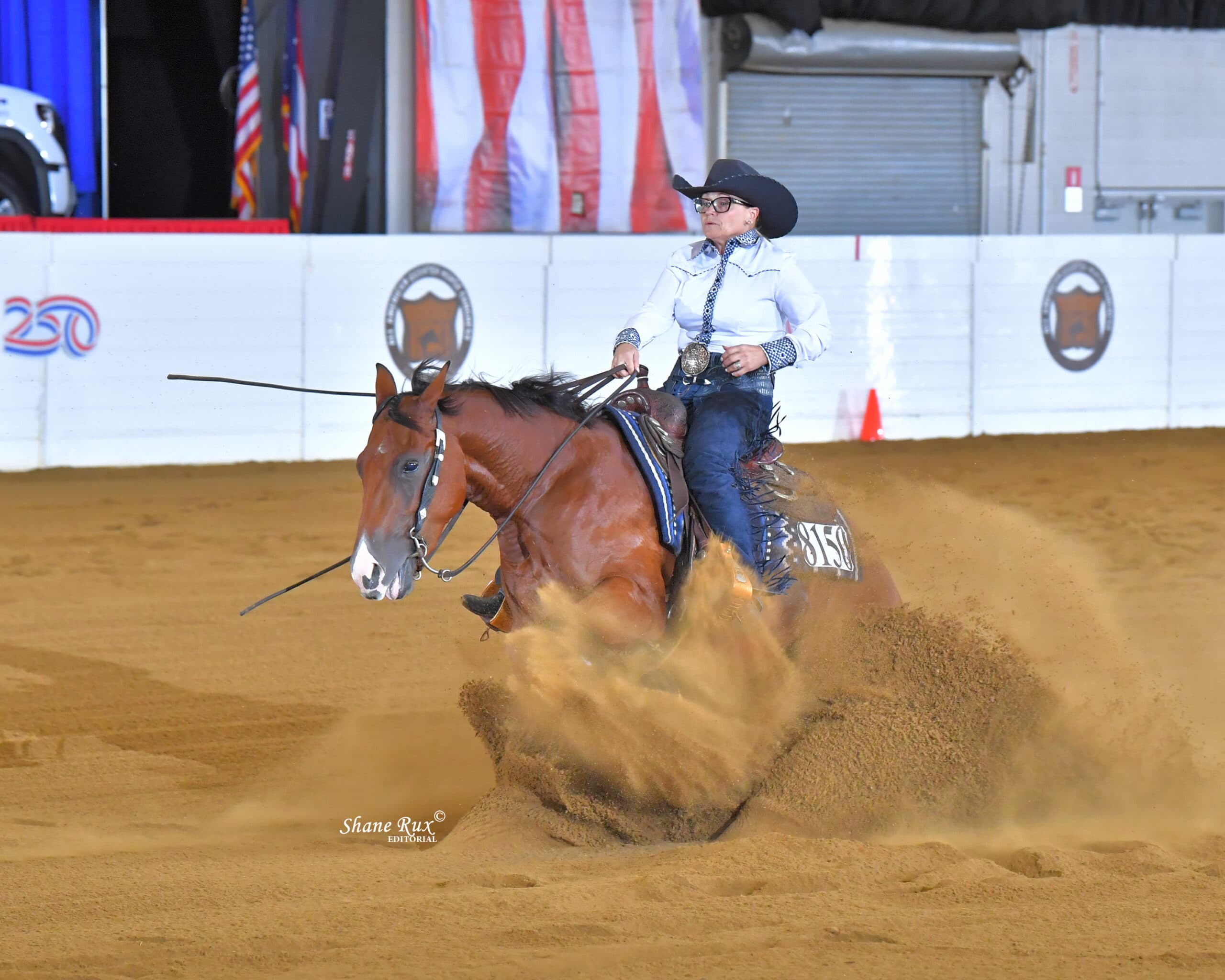 Cowgirl in white shirt and blue jeans rides a bay horse at speed through dirt arena during Western horsemanship competition, kicking up dust