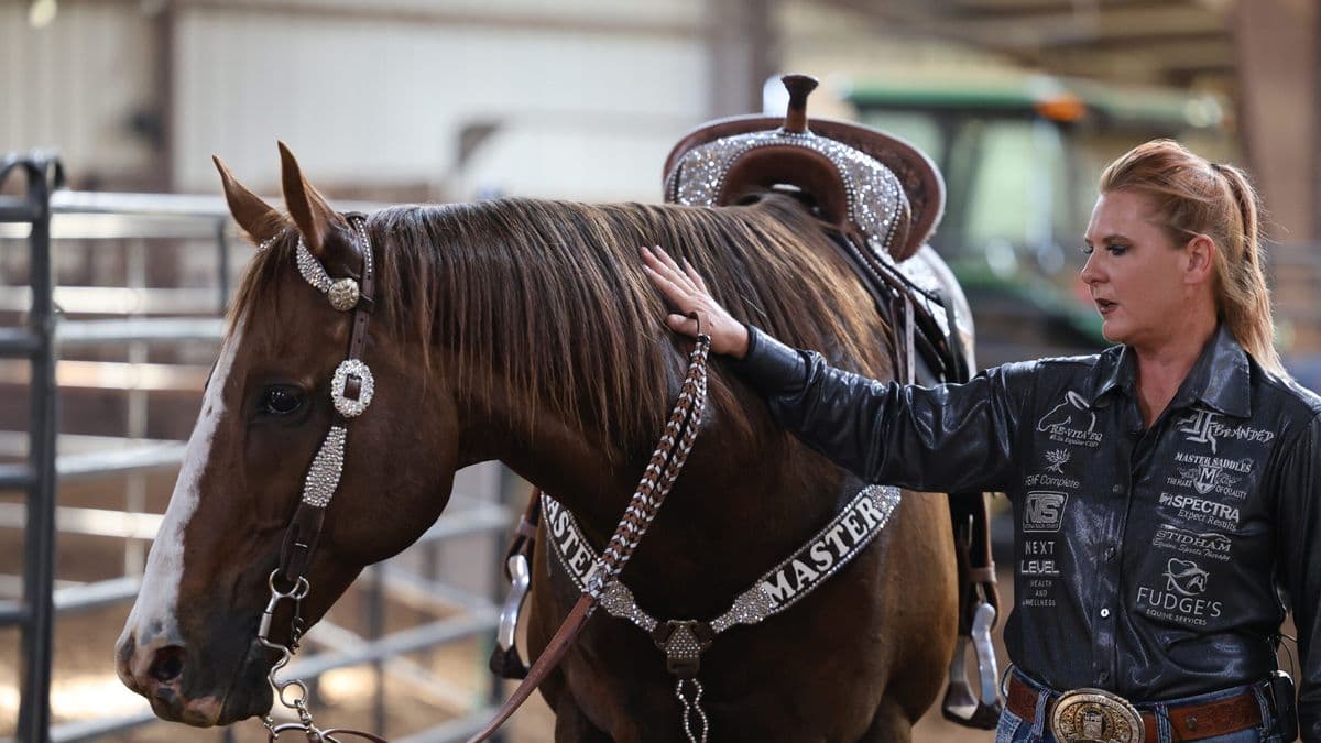 Woman in a sponsor-covered black shirt pats the neck of a sorrel horse wearing a rhinestone headstall and breast collar