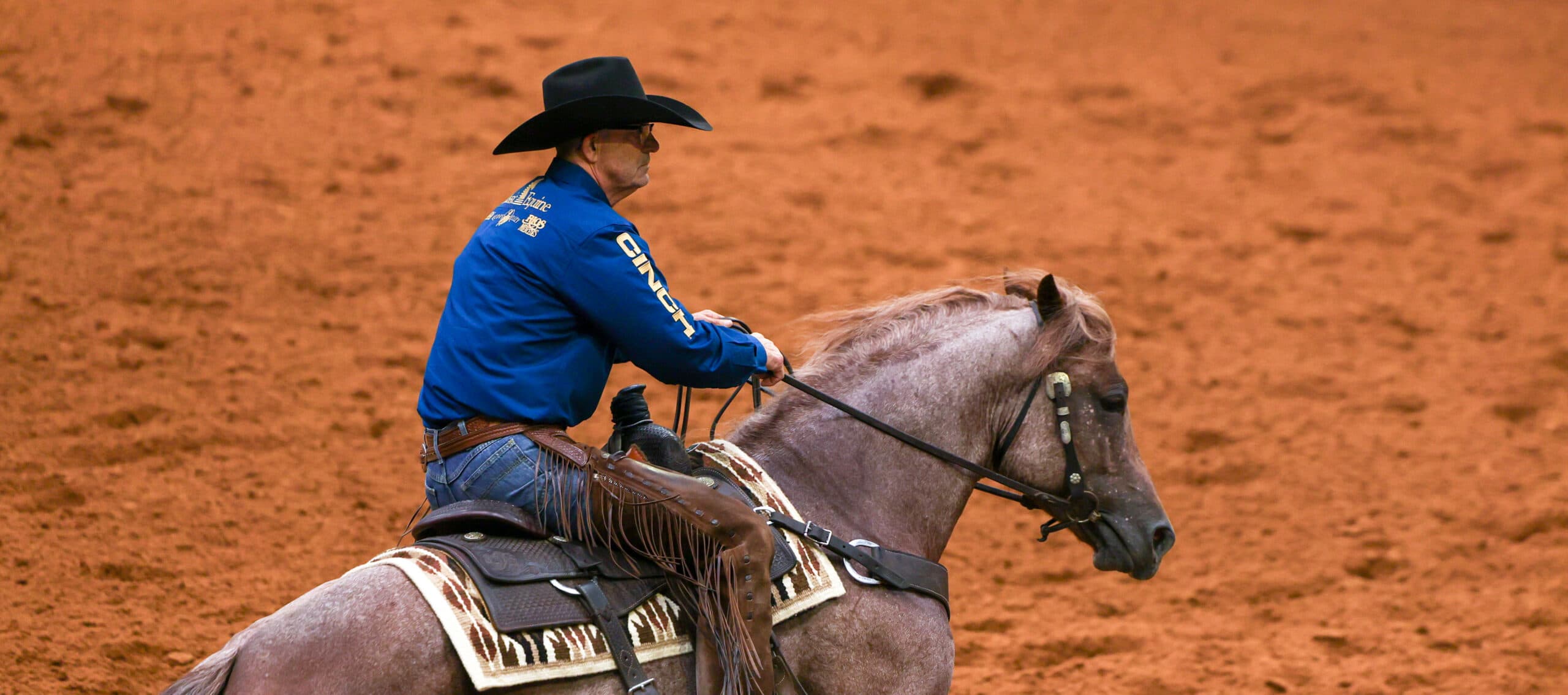Cowboy in blue shirt and black hat rides gray horse across red dirt arena during rodeo event