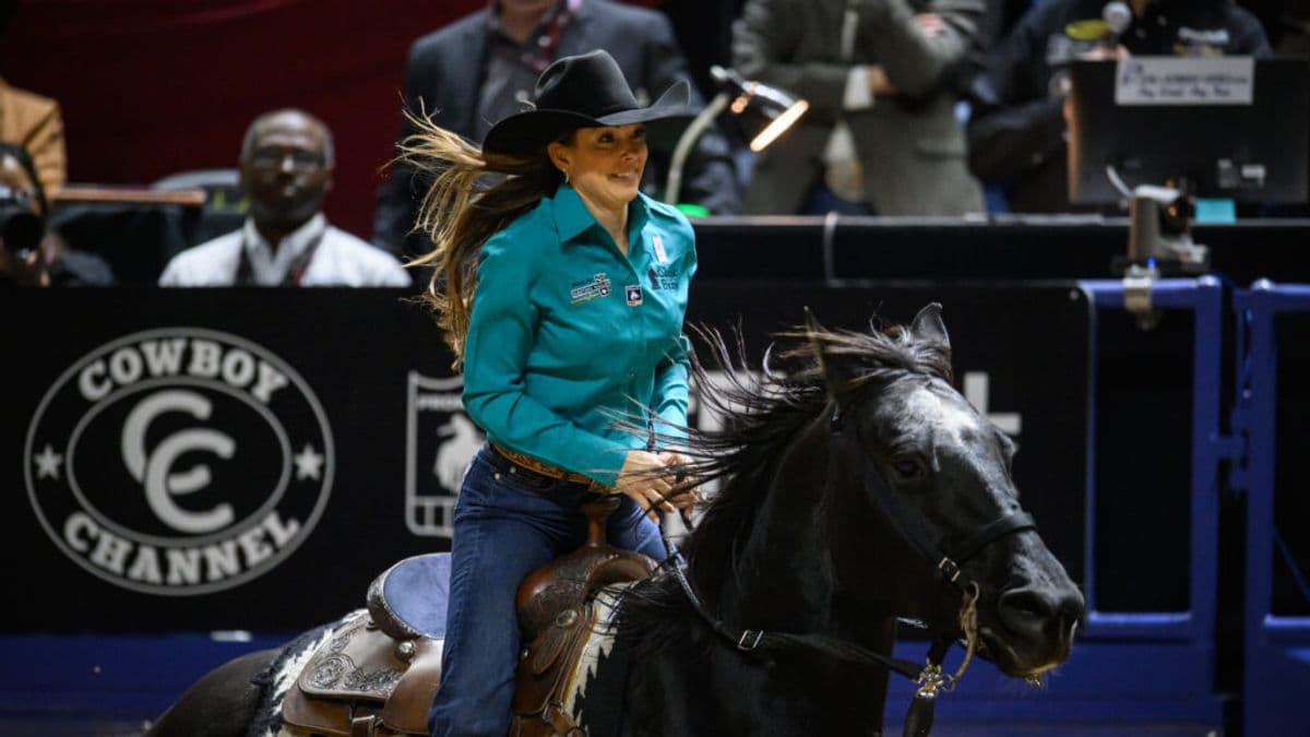 Smiling cowgirl in a teal shirt and black hat rides a dark horse past the Cowboy Channel arena banner