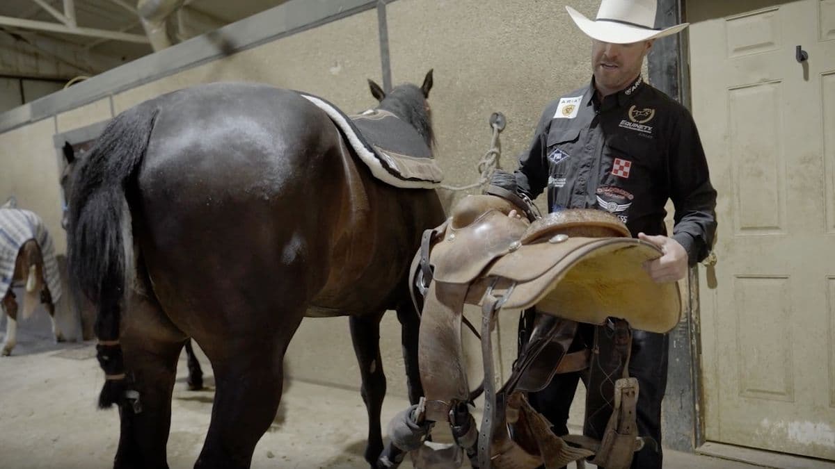 Cowboy in a white straw hat and sponsor shirt lifts a western roping saddle toward a dark horse in a barn aisle