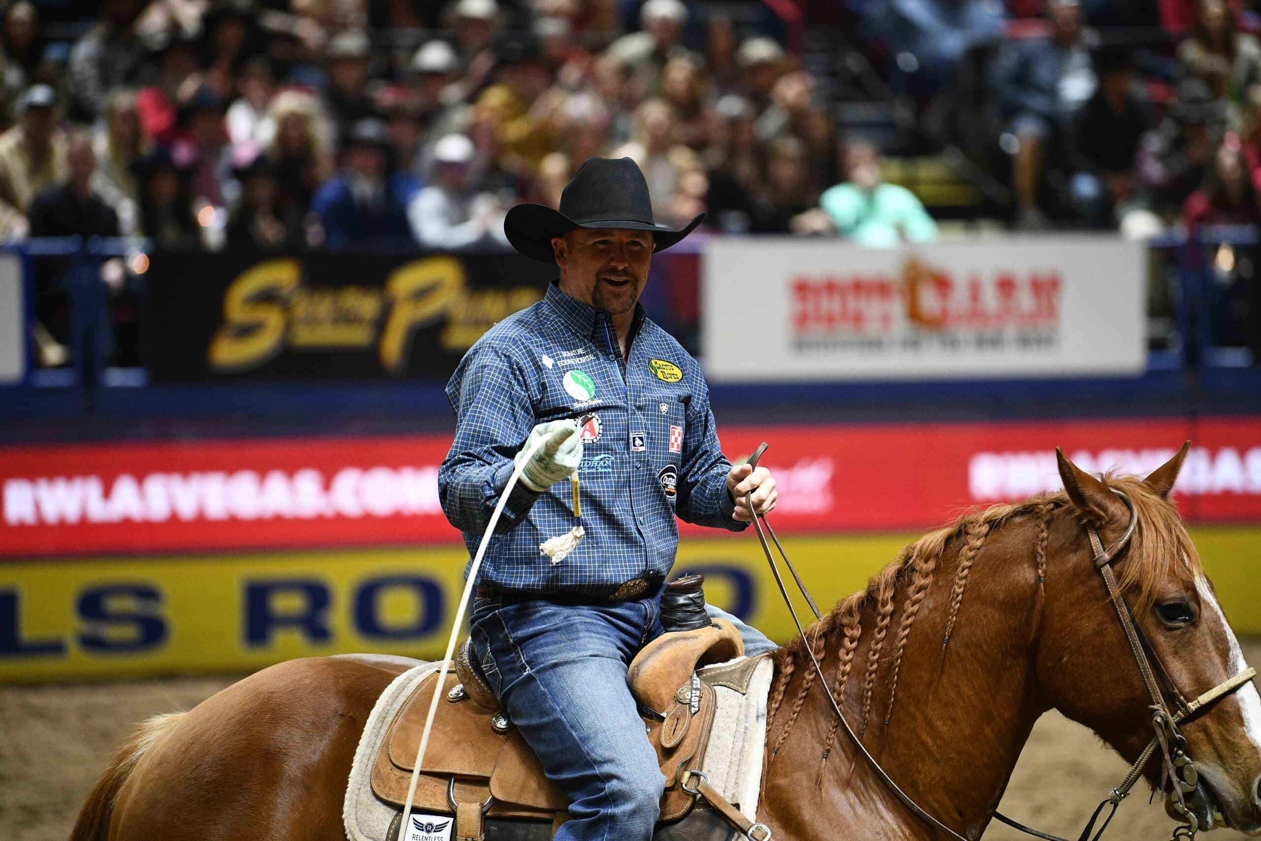 Cowboy on horseback wearing a blue checkered shirt and black cowboy hat rides in a rodeo arena before a crowd of spectators