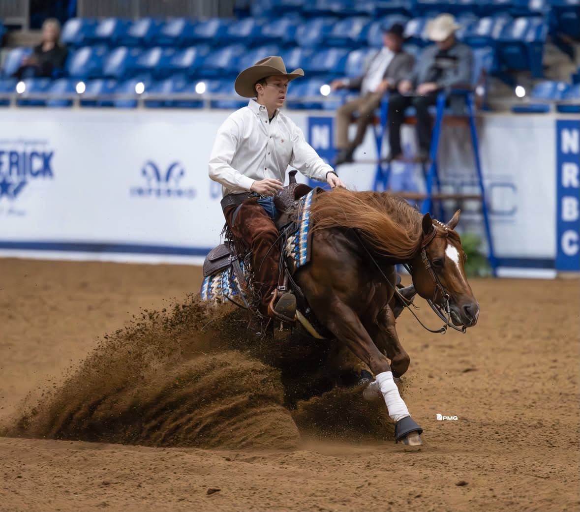 Barrel racer on brown horse kicks up dirt while making a turn at a rodeo arena with blue seating in background