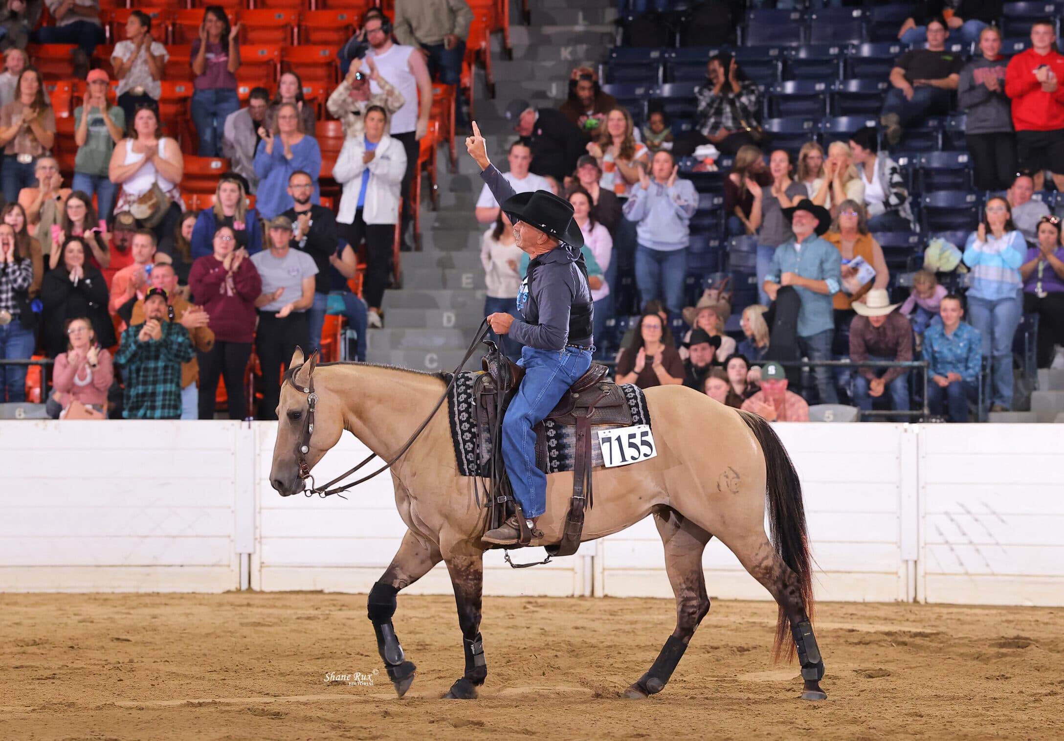 Rider on tan horse with number 7155 acknowledges crowd during rodeo event as spectators in bleachers applaud