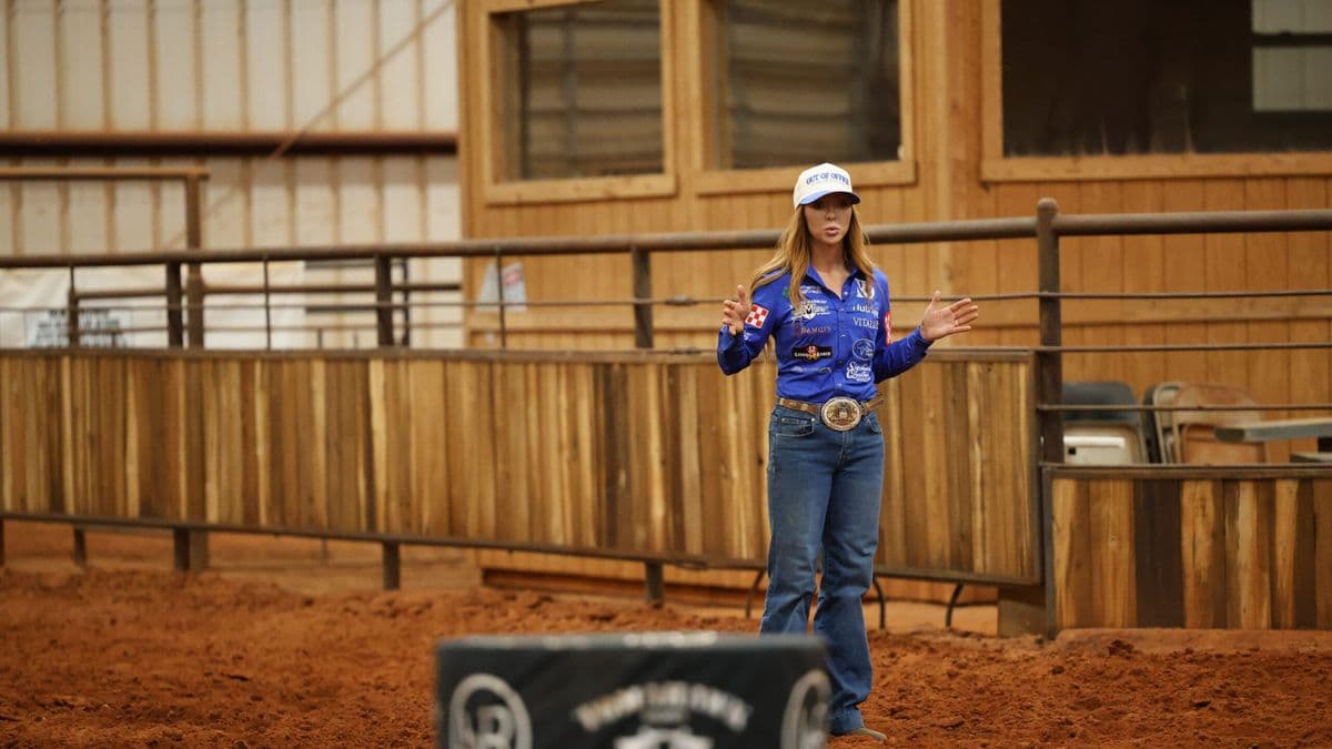 Woman in a blue sponsor shirt and ball cap gestures with both hands while speaking in a dirt arena beside a padded barrel