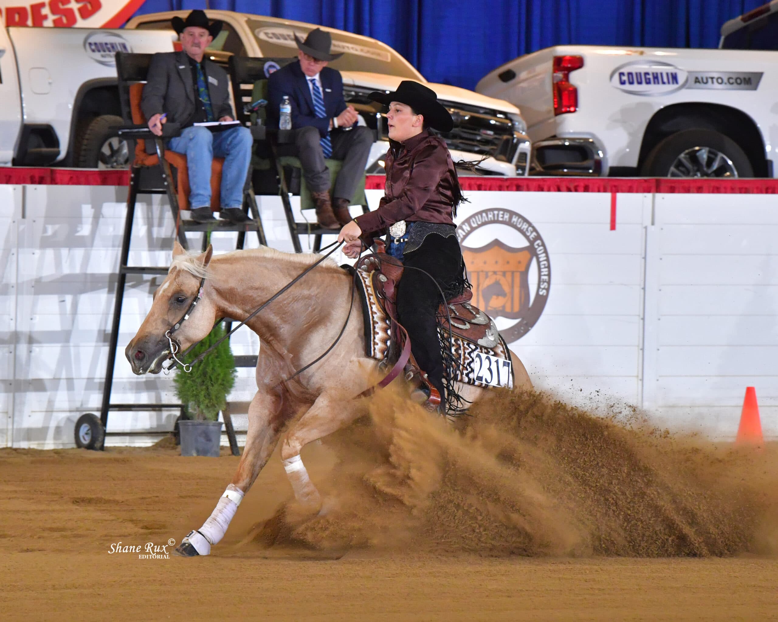 Barrel racer on palomino horse kicking up dust while turning around barrel at indoor rodeo arena with judges watching from elevated stand