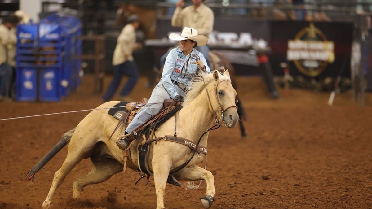 Rodeo competitor in light blue shirt and cowboy hat rides a palomino horse while roping during an indoor rodeo event
