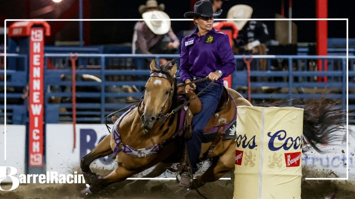 Barrel racer on brown horse in purple jacket and black hat rounds a barrel during nighttime rodeo competition, with Coors signage visible