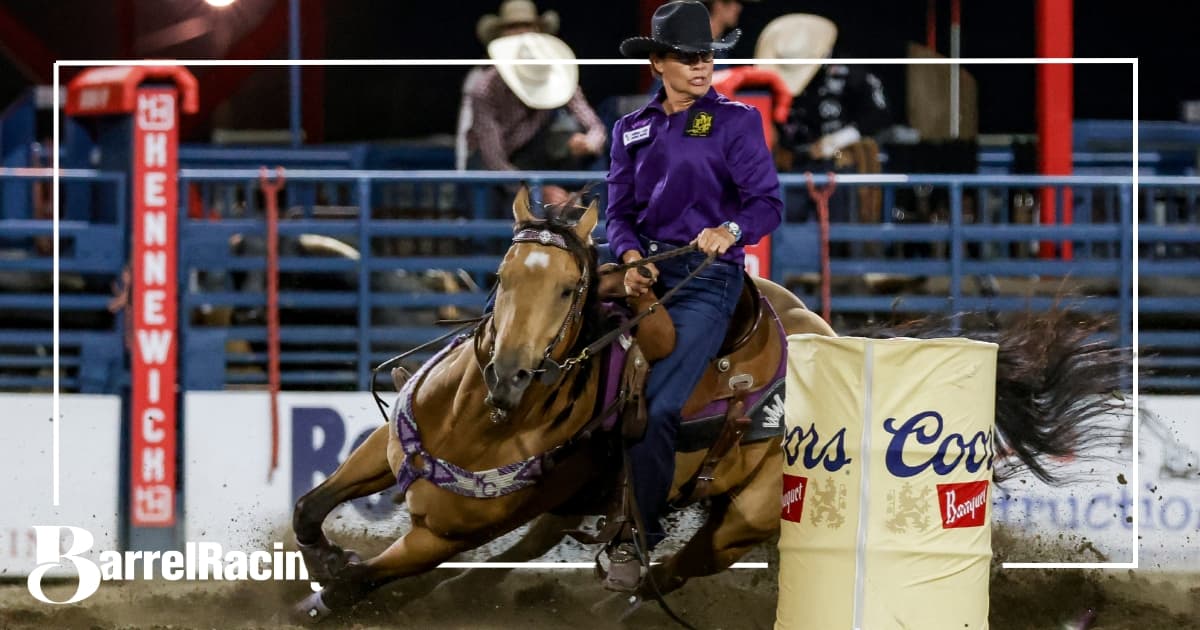 Barrel racer on brown horse in purple jacket and black hat rounds a barrel during nighttime rodeo competition, with Coors signage visible