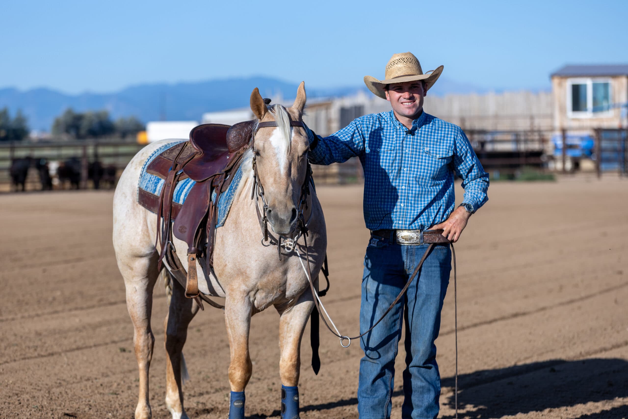 Cowboy in blue shirt and tan hat leads a saddled horse across a dirt arena with mountains in the background