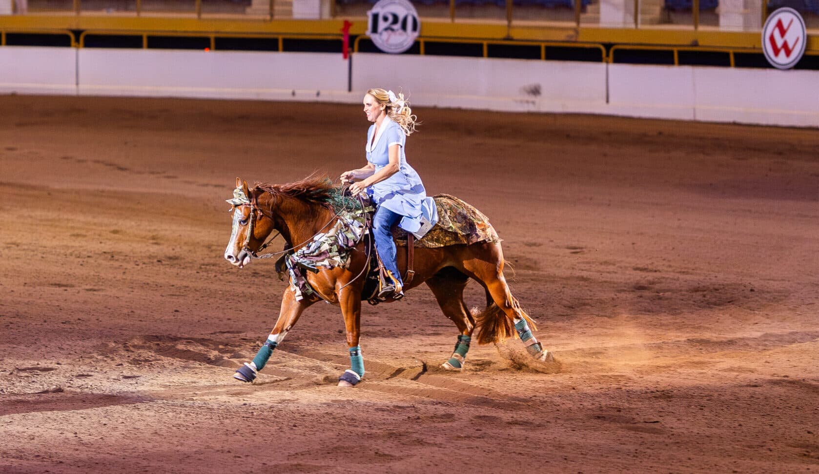 Barrel racing competitor in blue shirt rounds a barrel on a brown horse at a rodeo arena with sponsor signage visible