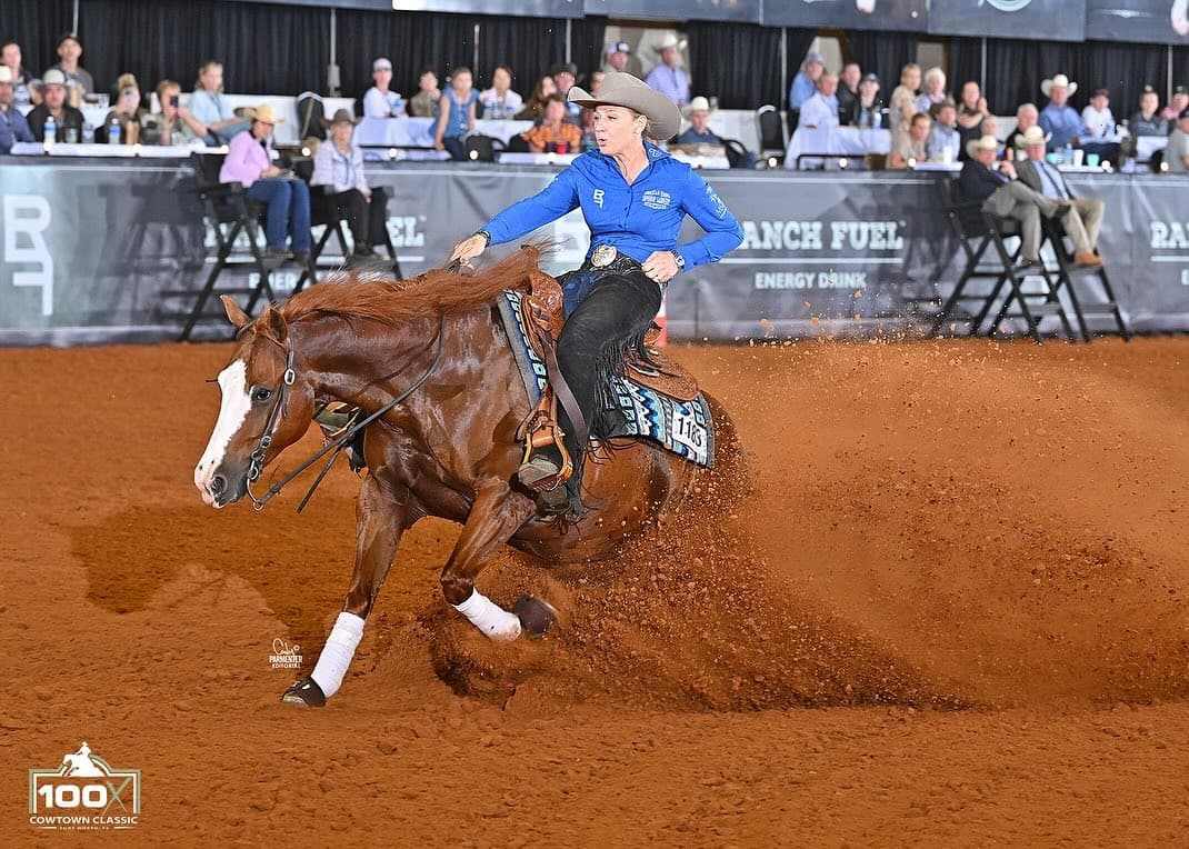 Reining rider in a blue shirt and cowboy hat slides her sorrel horse to a stop, spraying dirt, at the Cowtown Classic
