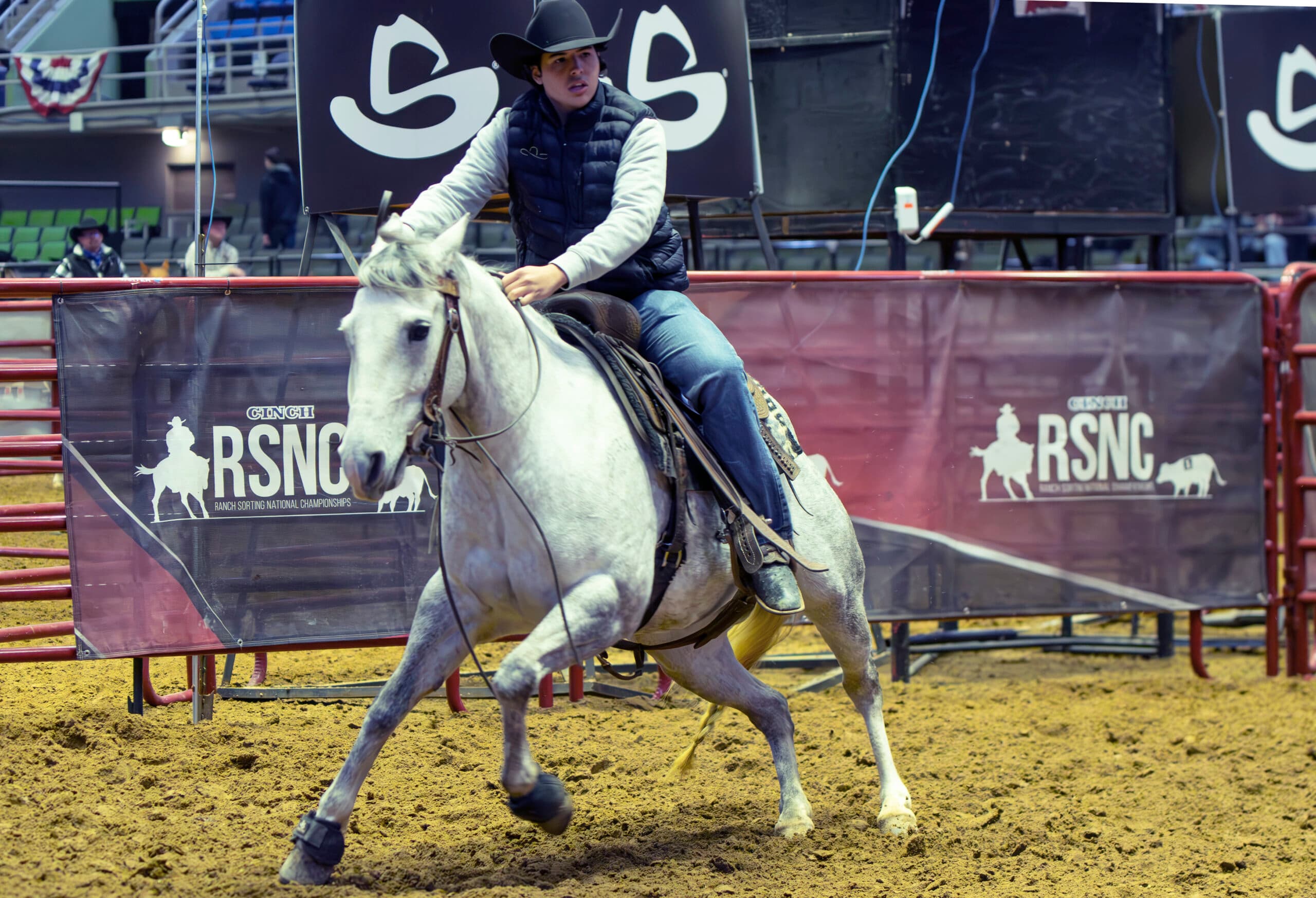 Rider in black cowboy hat and puffy vest turns a gray horse in the arena dirt past Cinch RSNC banners