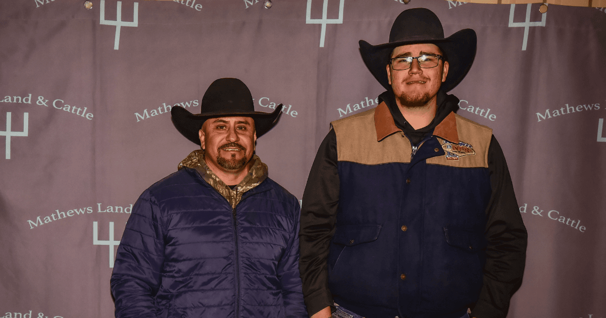 Two cowboys in black hats and winter jackets pose side by side in front of a Mathews Land & Cattle backdrop