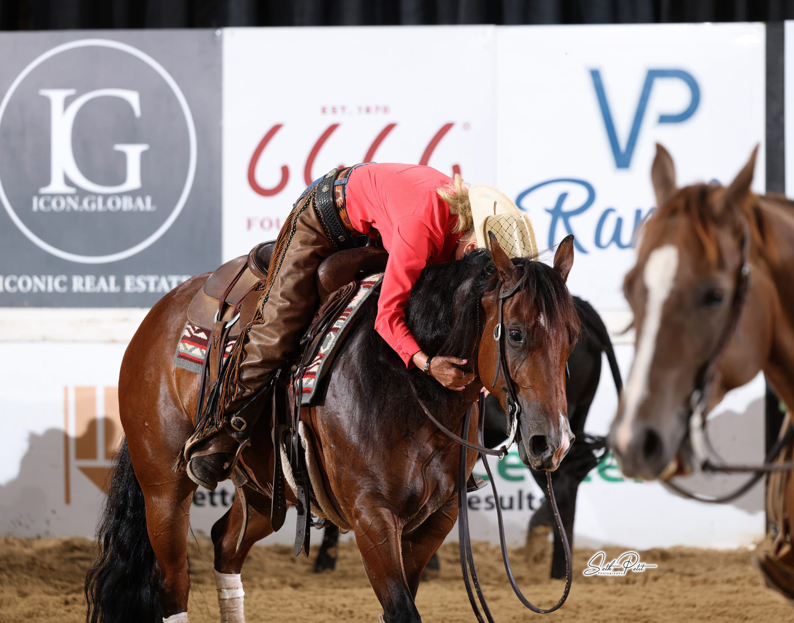 Rider in a red shirt and straw hat leans forward to hug her bay horse's neck in a sandy arena after a run