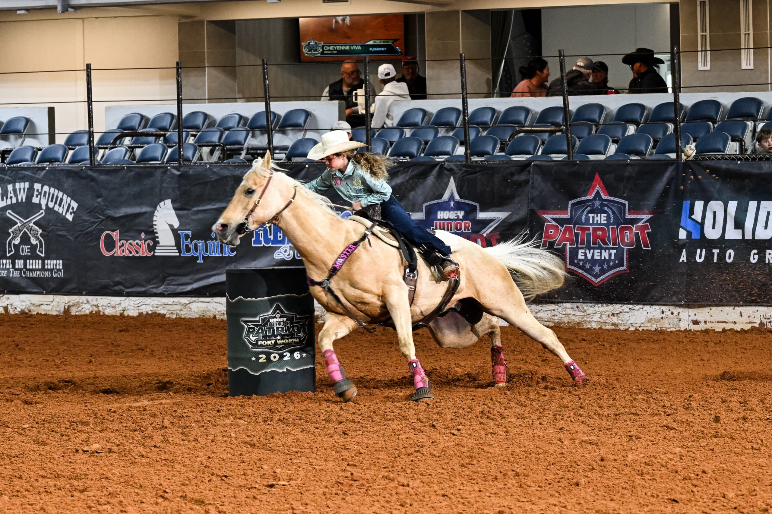 Young barrel racer in a straw cowboy hat rounds a Hooey Junior Patriot barrel on a palomino horse in pink splint boots