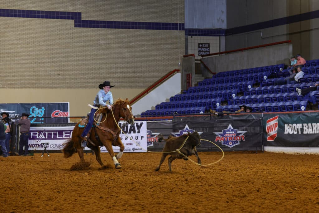 Breakaway roper on a sorrel horse swings her loop toward a running calf in a dirt arena with sponsor banners behind