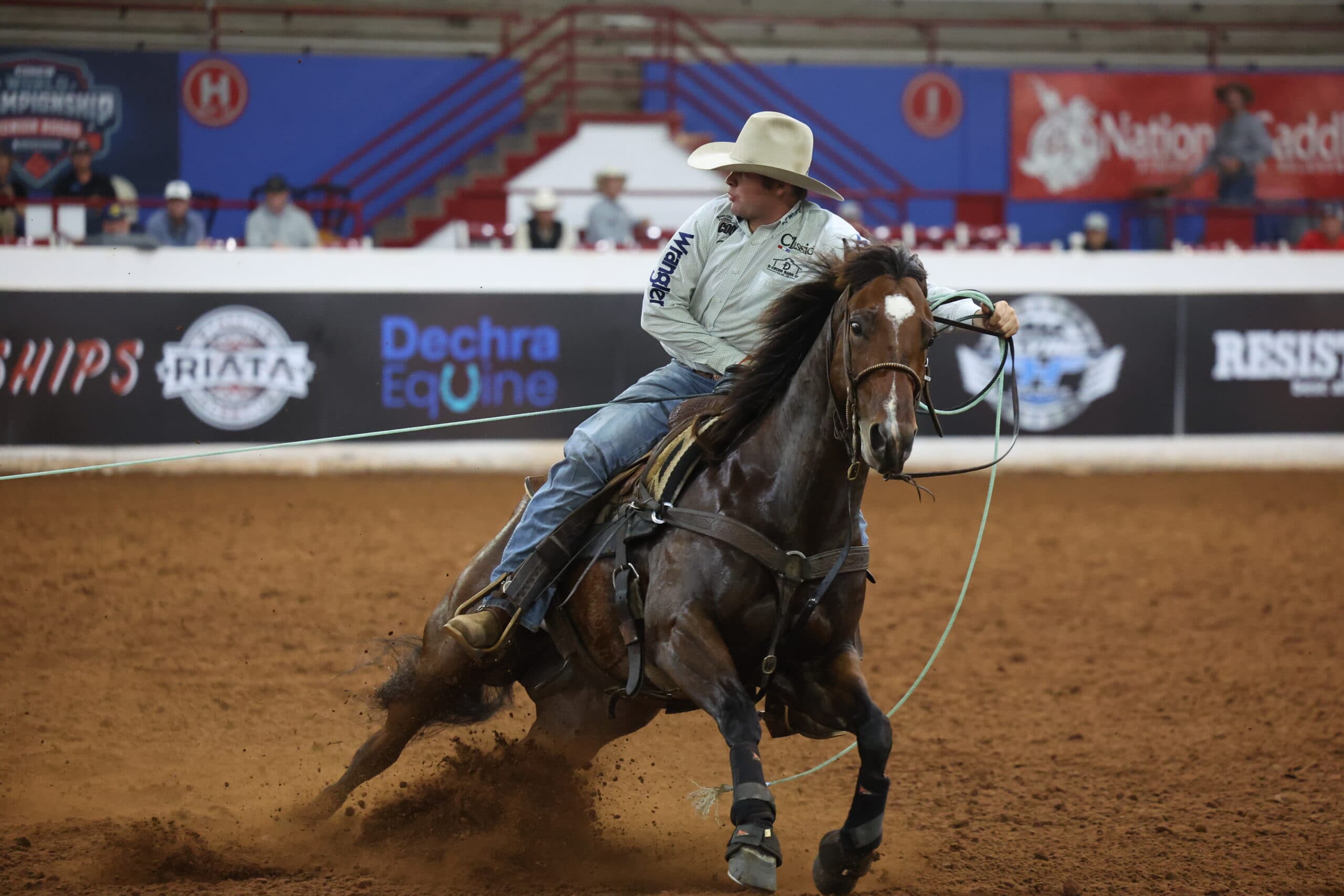 Roper on brown horse executing a breakaway roping run, sliding through dirt arena with rope extended during indoor rodeo competition