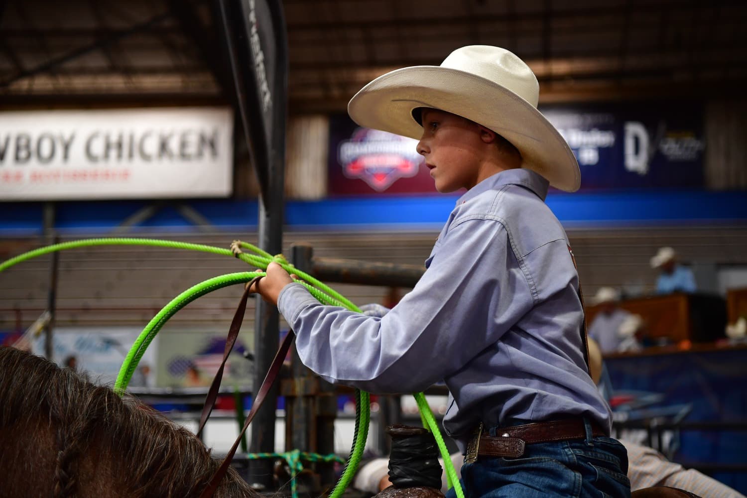 Young roper in light blue shirt and cream cowboy hat holds a bright green rope while mounted, with indoor arena and "Cowboy Chicken" signage visible in background