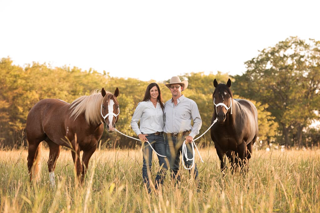 Man and woman in western attire holding ropes stand between two saddled horses in a pasture
