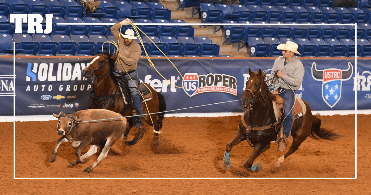 Two ropers on horseback work together during team roping competition at an indoor rodeo arena with sponsorship signage visible