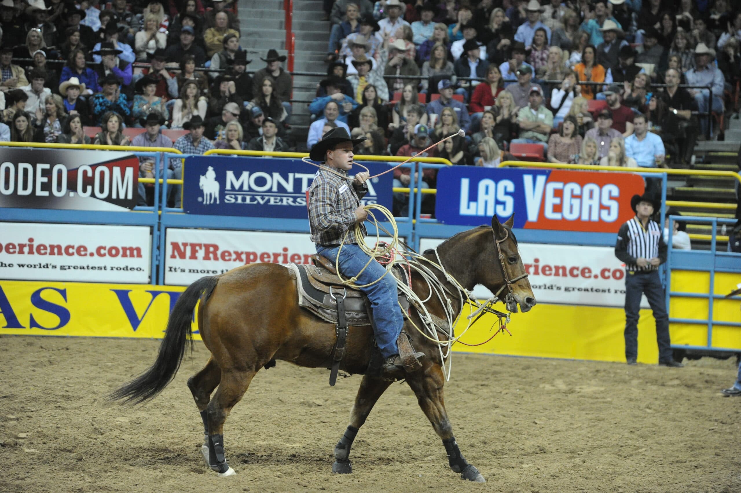 Cowboy in a black hat swings his rope while riding a bay horse across the arena dirt at a packed indoor rodeo