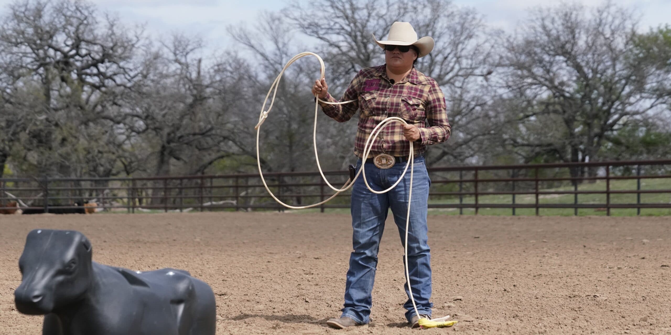 Roper in a cowboy hat, plaid shirt and trophy buckle swings a loop on foot toward a plastic roping dummy steer in a dirt arena
