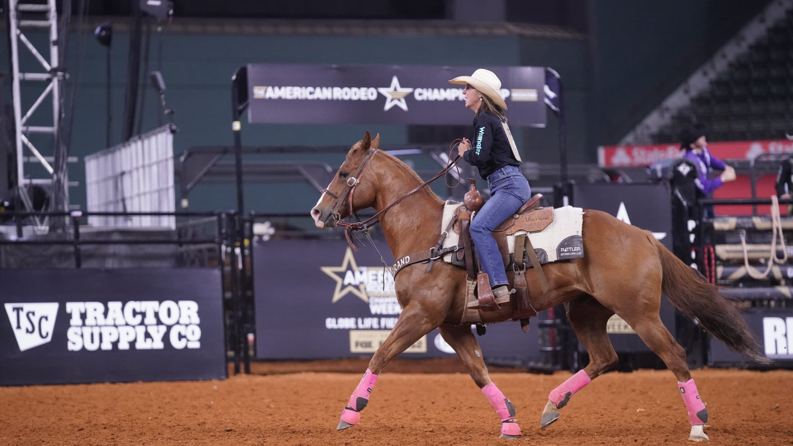 Breakaway roper in a straw cowboy hat loping her sorrel horse in pink splint boots across the arena at The American Rodeo