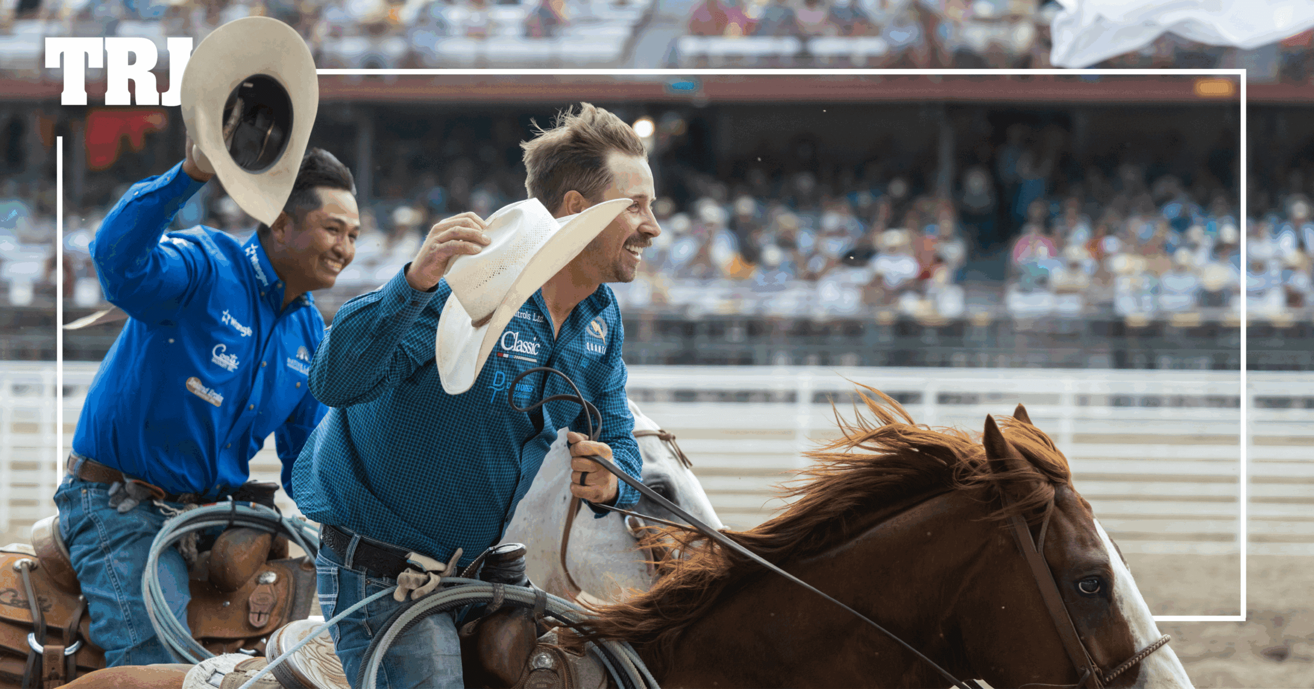 Cowboy on horseback waves his hat to the crowd at a rodeo arena while a fellow competitor in blue shirts rides alongside him