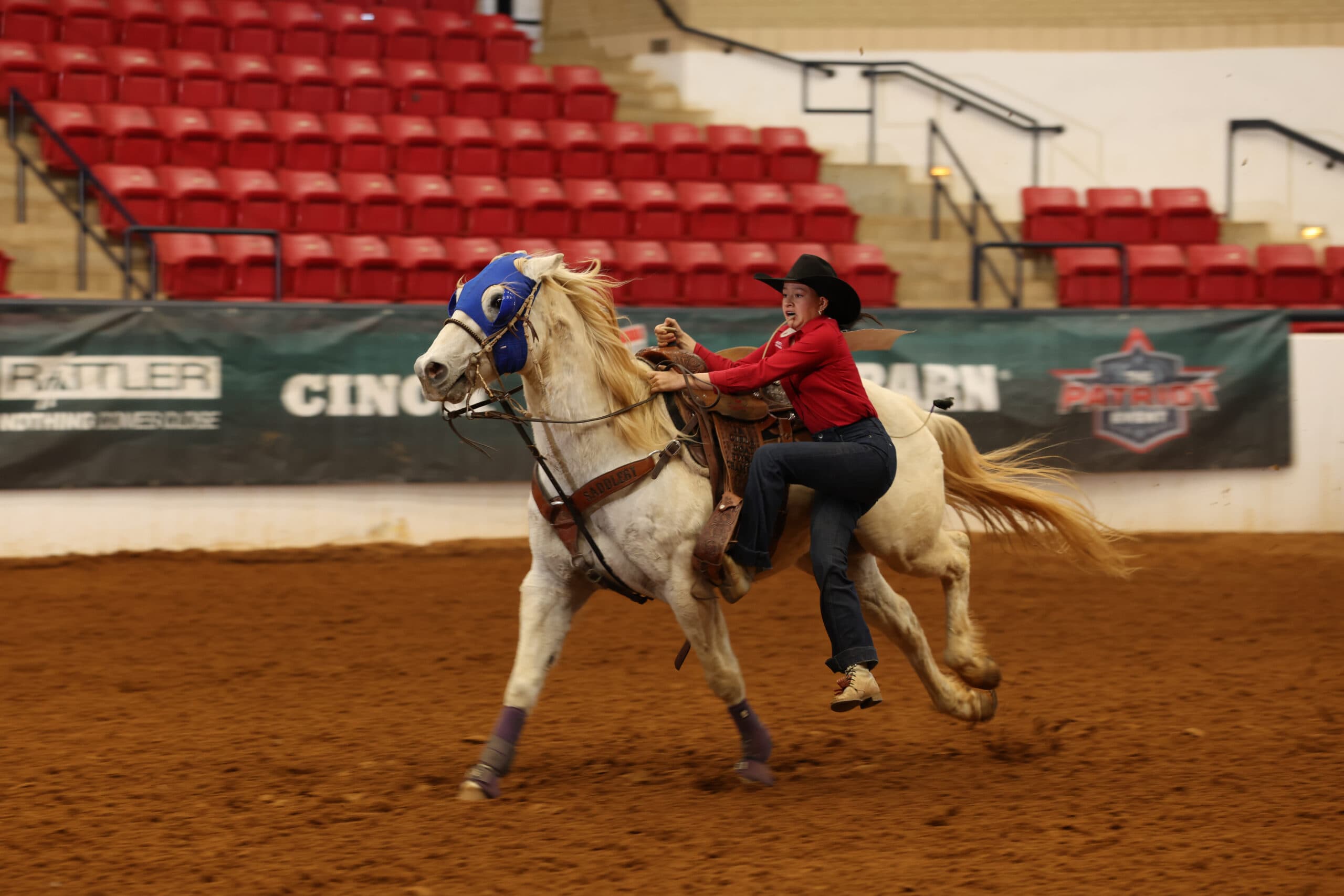 Barrel racer in a red shirt and black cowboy hat gallops a palomino horse wearing a blue hood across the arena dirt
