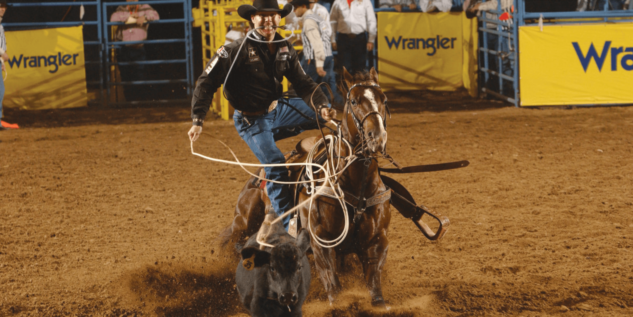 Tie-down roper dismounting his sorrel horse with rope in teeth as the roped calf runs ahead in a dusty rodeo arena
