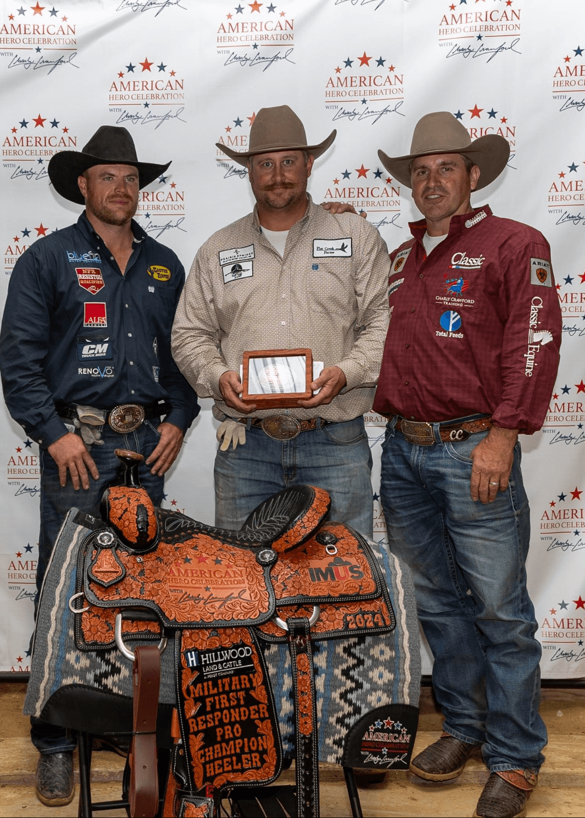 Three cowboys in hats stand with a trophy plaque behind a tooled champion heeler saddle at an American Hero Celebration backdrop