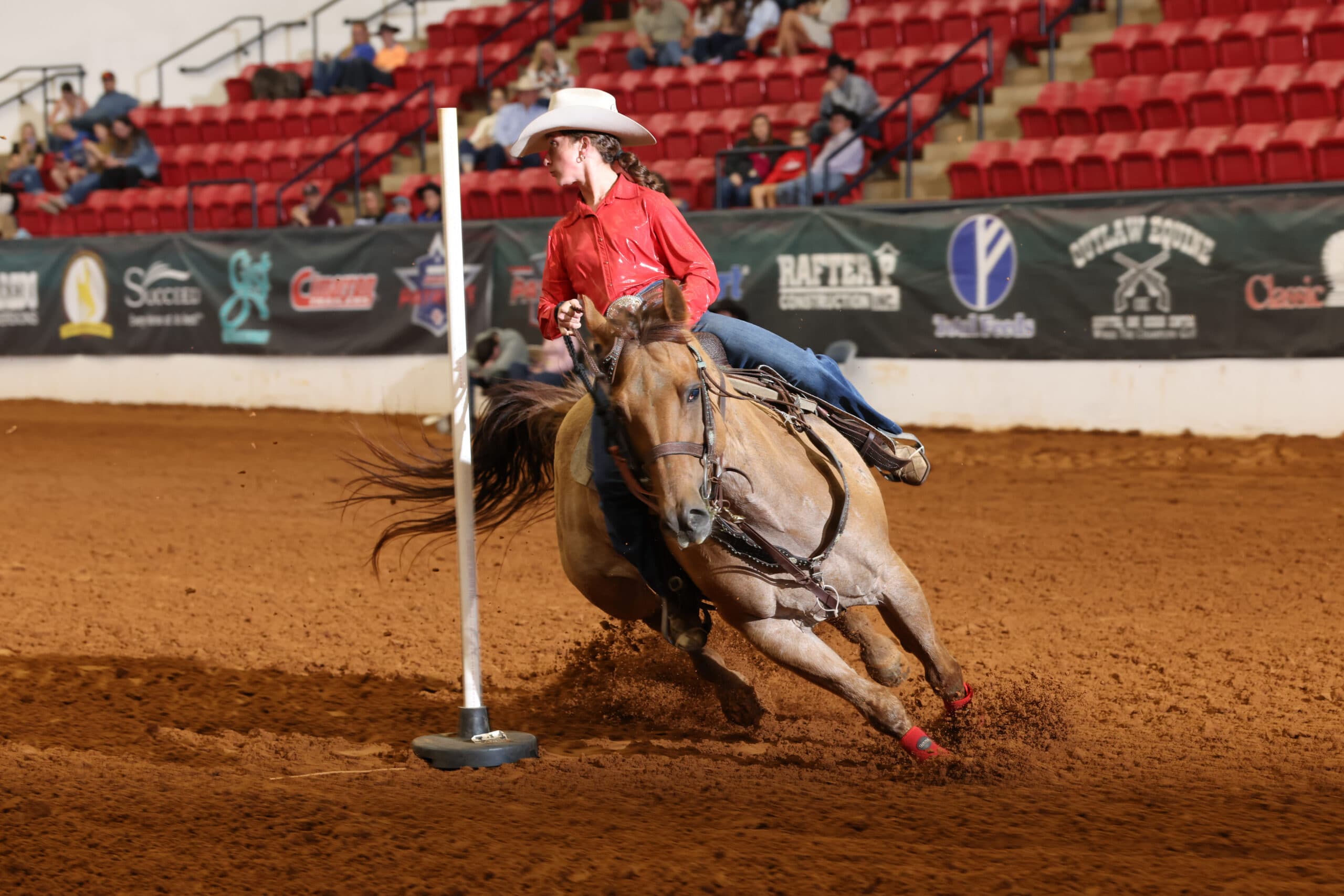 Barrel racer in a red shirt and white cowboy hat rounds a pole on a dun horse in red leg wraps, kicking up arena dirt