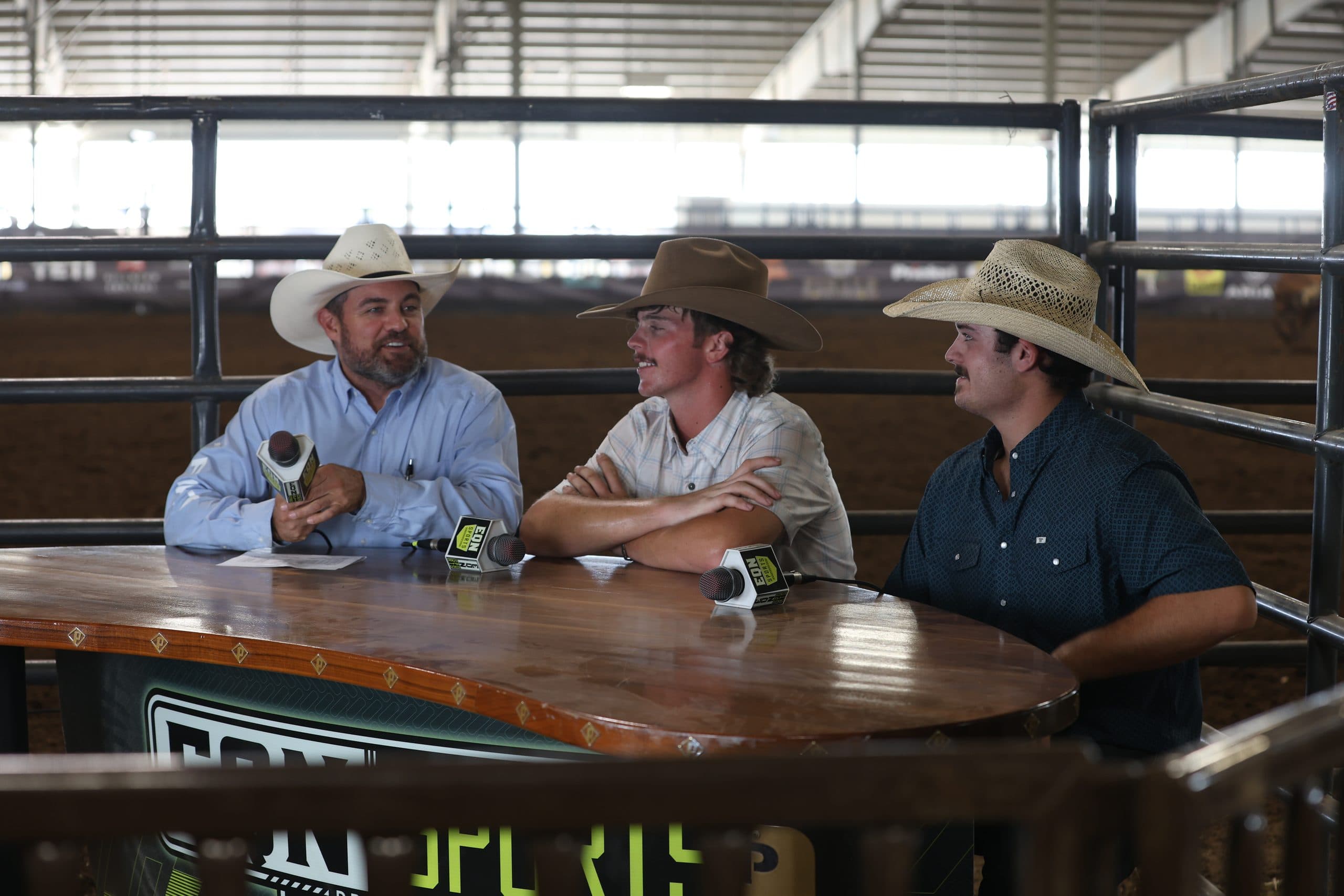 Three cowboys in cowboy hats sit at a table during what appears to be a rodeo broadcast or interview at an indoor arena