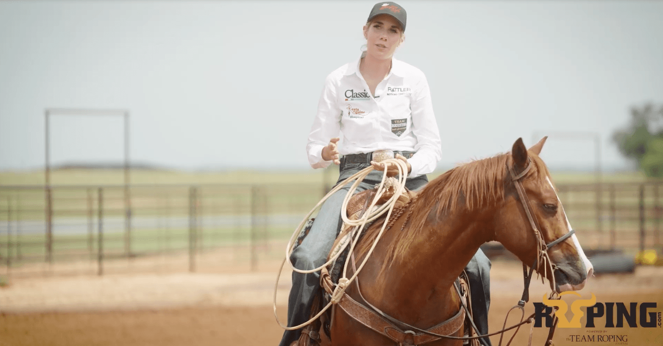 Woman in a white sponsor shirt and cap sits on a sorrel horse holding a coiled rope, gesturing as she explains roping technique