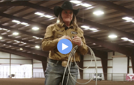 Woman in a black cowboy hat and tan western shirt holding a coiled rope in an indoor arena, video play button overlaid