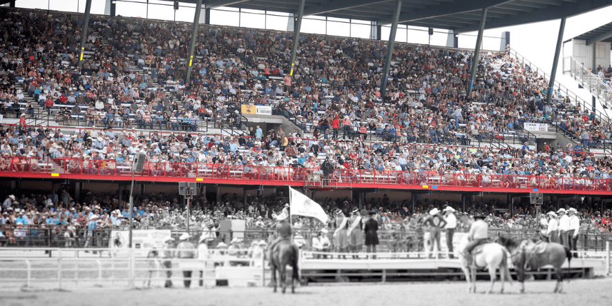 Mounted riders carrying flags line up in a rodeo arena before a packed grandstand of spectators
