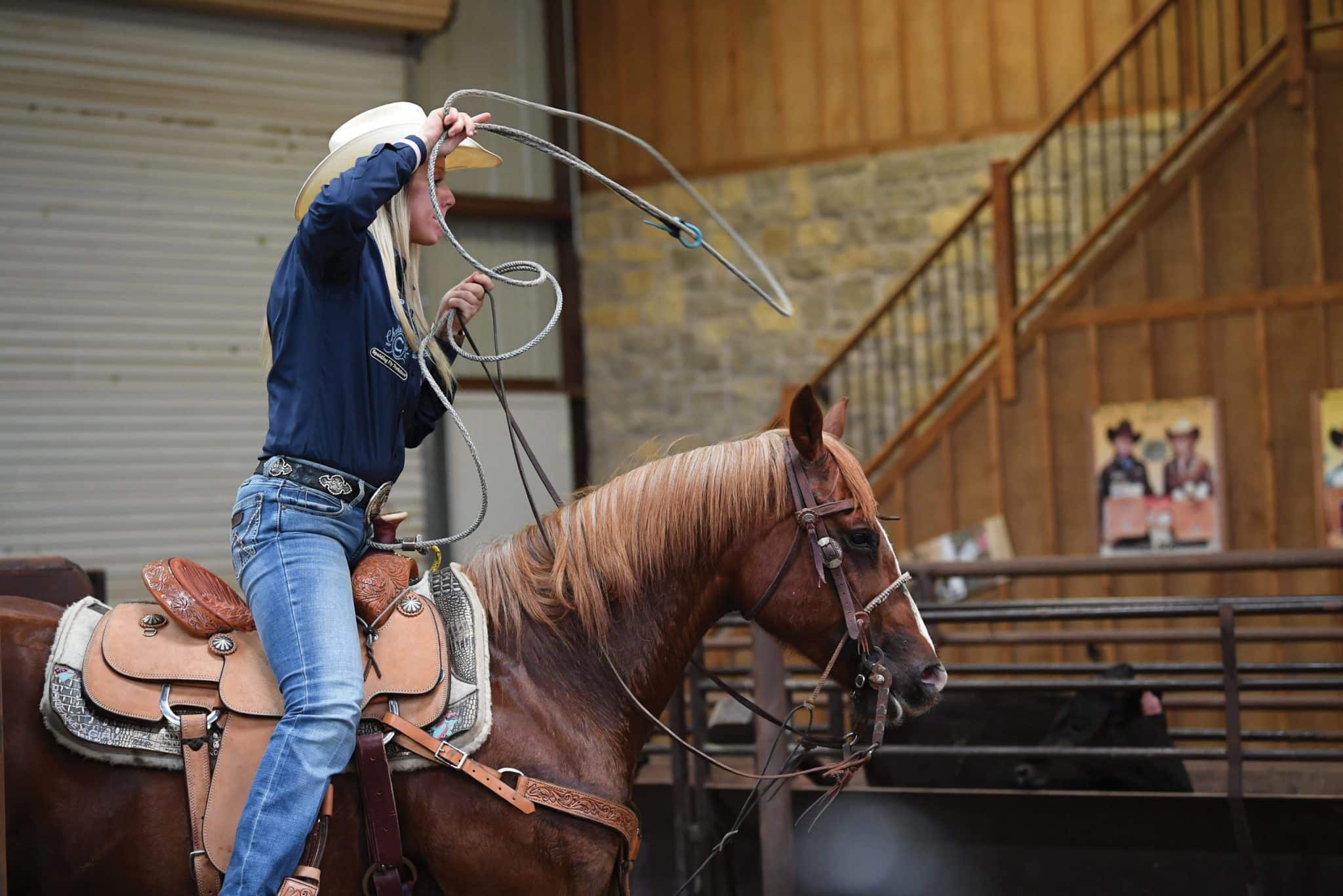 Blonde cowgirl in a navy shirt and white hat swings a rope loop overhead while mounted on a sorrel horse in an indoor arena
