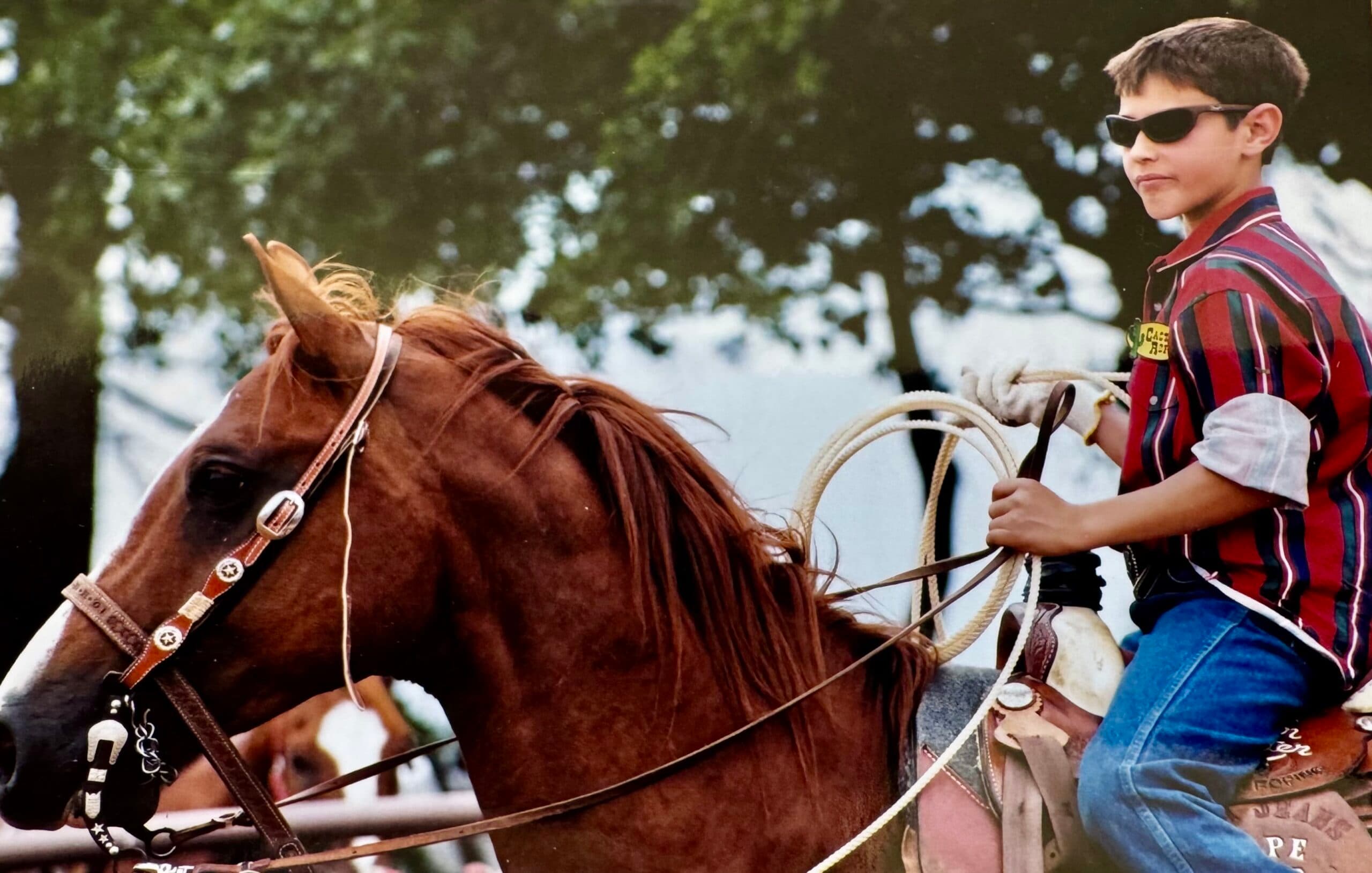 Young roper in sunglasses and striped shirt holds a coiled rope while riding a sorrel horse at a rodeo