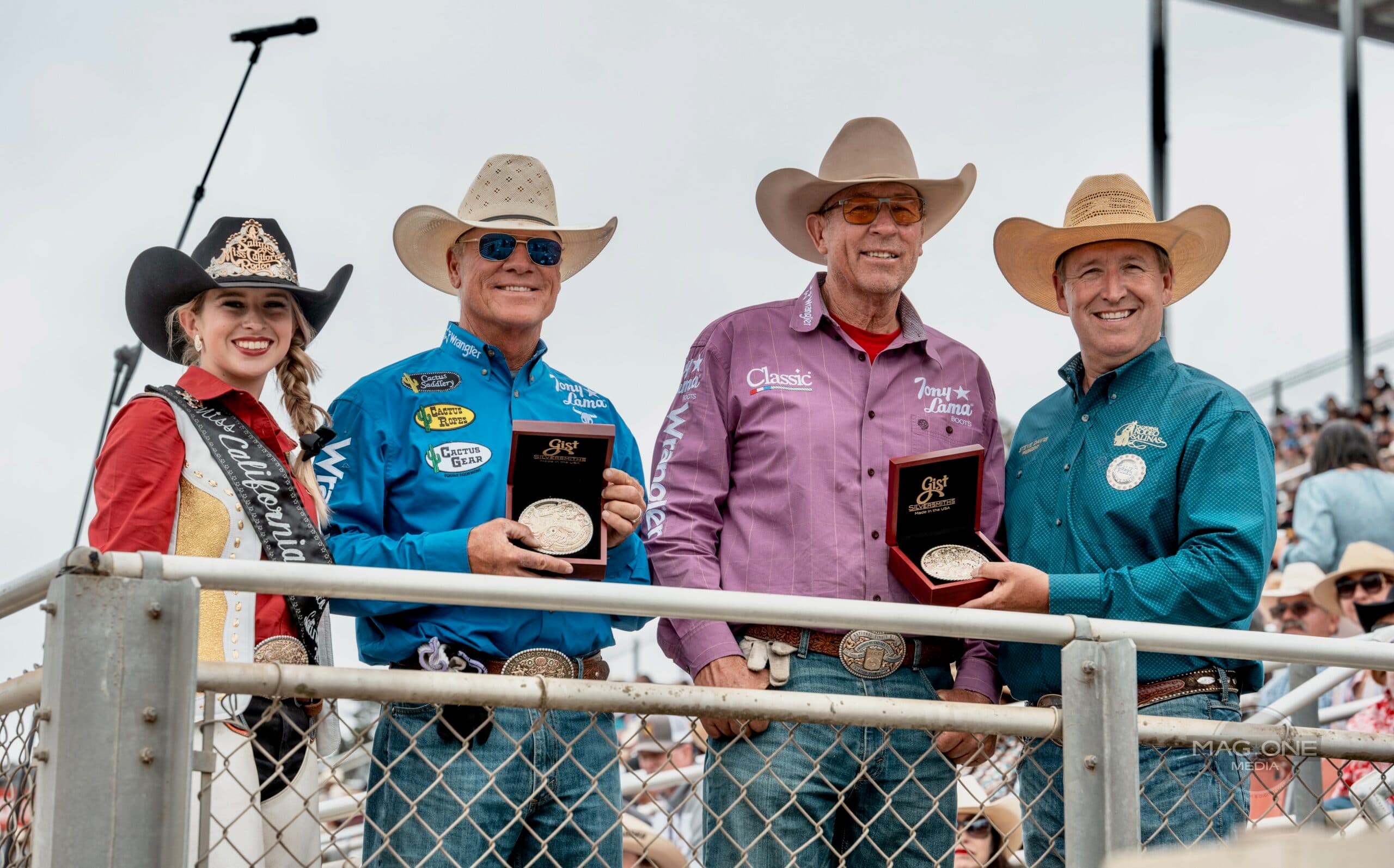Four rodeo competitors in western attire hold awards at a stadium event, with a woman on the left holding a microphone and three men displaying buckles and trophies