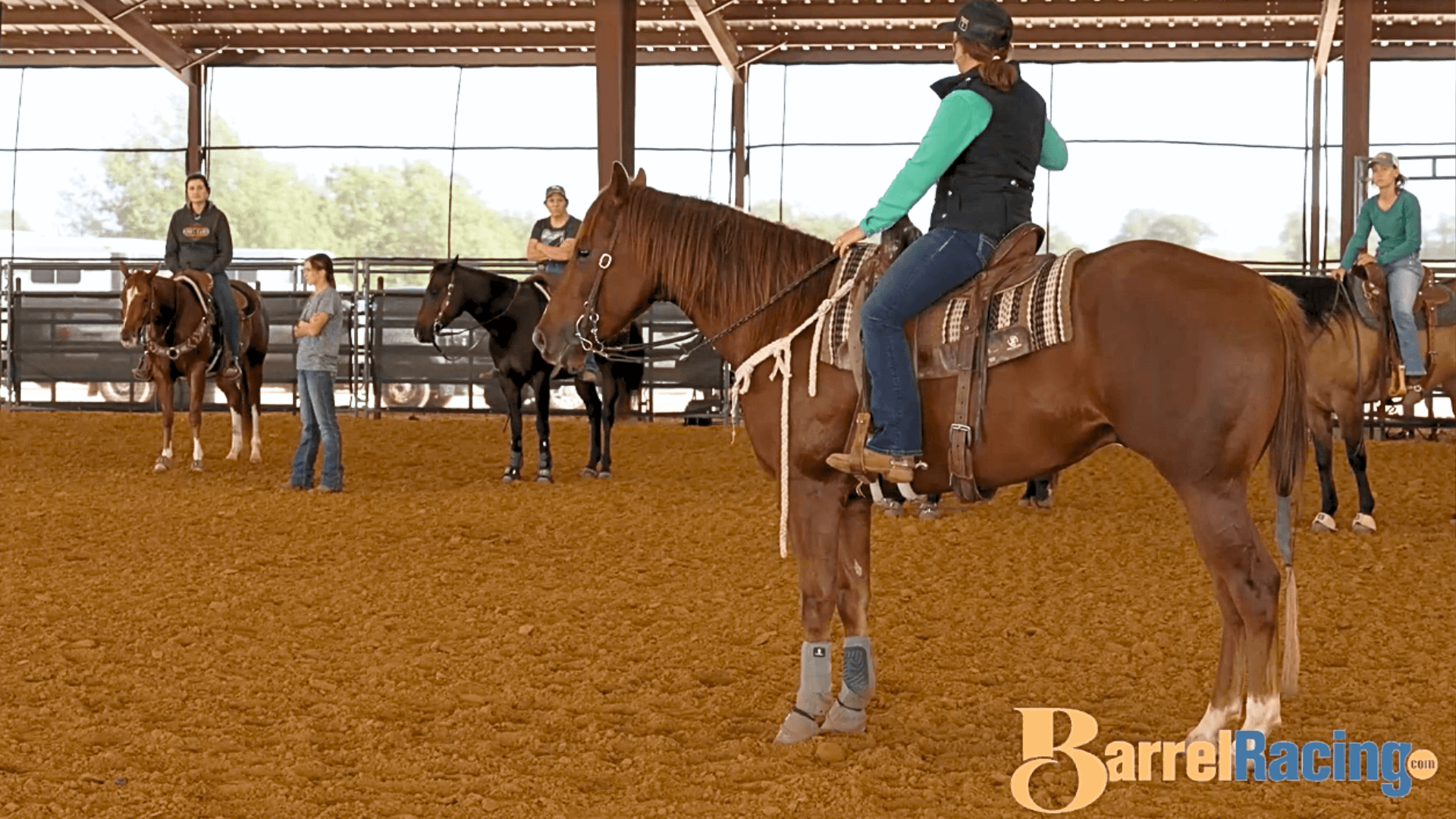 Rider in a green shirt and black vest sits on a sorrel horse in a covered arena as onlookers on horseback watch a barrel racing clinic